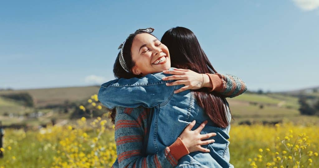 Two friends hug each other smiling in a field of yellow flowers.