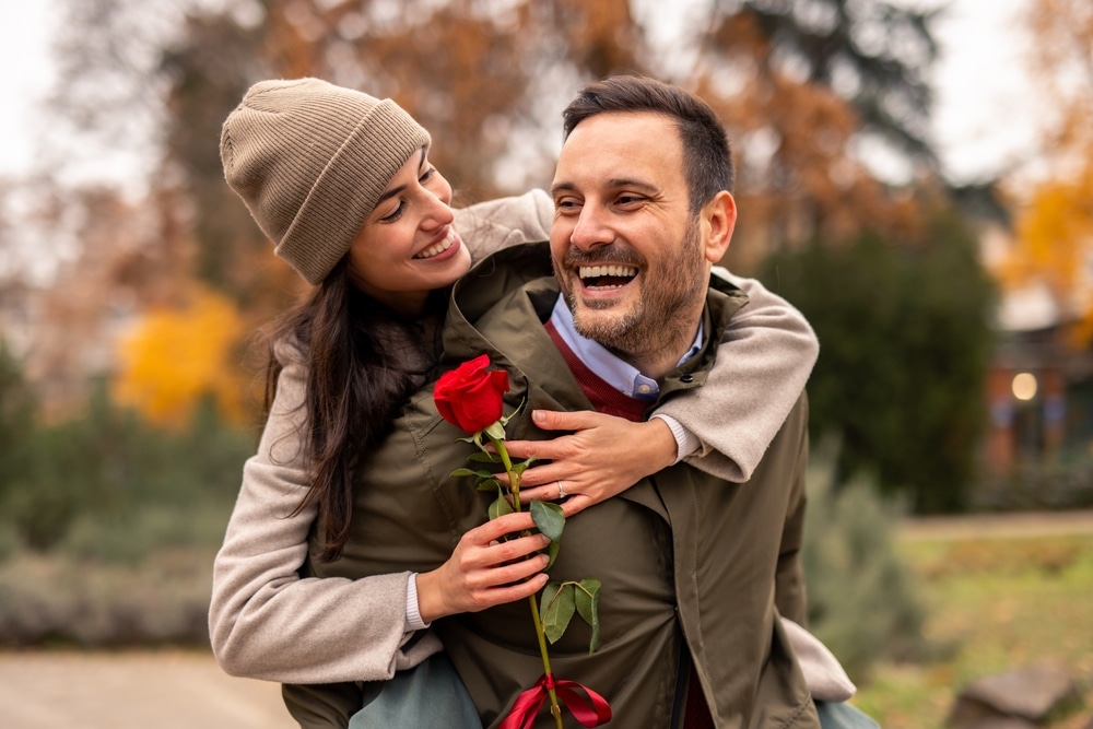 A man giving a woman a piggy back. She is holding a rose and they are both smiling