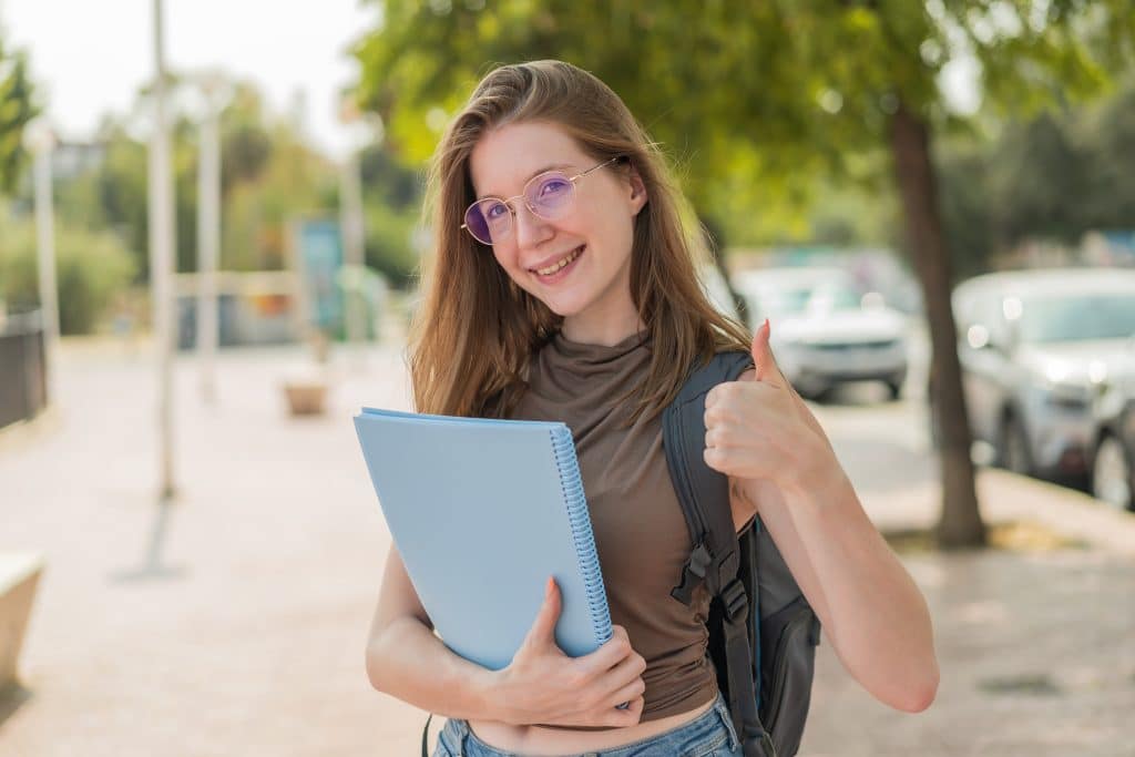 A smiling female student holding a folder and giving a thumbs up to the camera, on a journey to figure out her studies.
