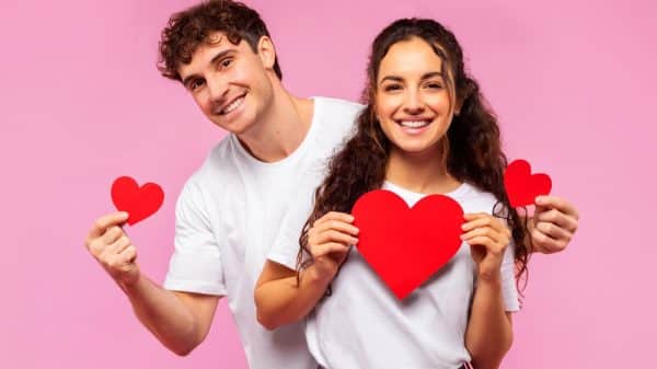 A boy and a girl holding red paper love hearts against a pink background.