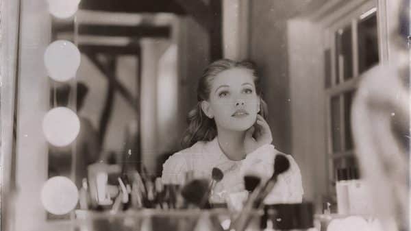 Black and white vintage photo of woman sitting at a makeup vanity with lots of makeup products in front of her and big lights around the mirror.