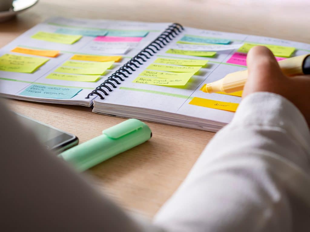 A hand with a highlighter hovering over a planner that is completely full of stick notes labeling jobs to do.