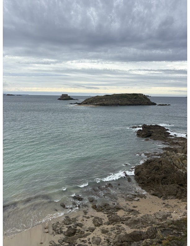 Picture of the coastline in Saint-Malo as seen from the city walls. Blue/green water with locks of rocks scattered on the sand.