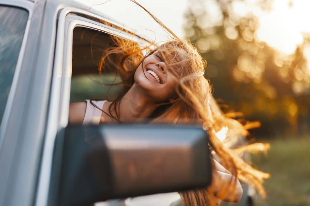 A woman, not caring about the push for perfection, leaning her head out of a car window and letting her hair blow in the wind.
