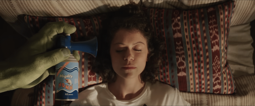 Tatiana Maslana as her character, She-Hulk, in She-Hulk: Attorney at Law. She is lying down, asleep, and a large green hand, Hulk, is holding an airhorn to her ear, about to fire it off.