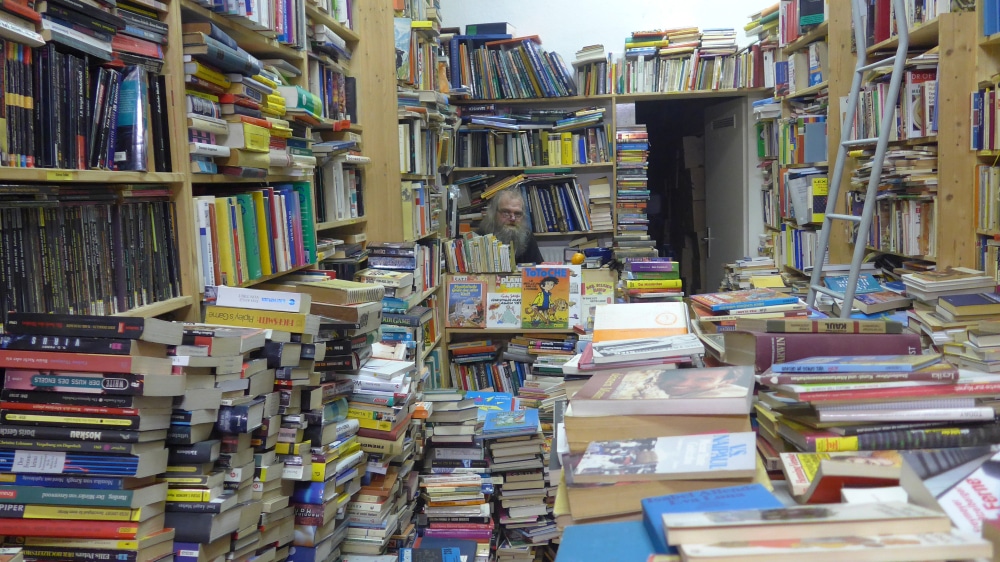 A man in a bookstore overflowing with many disorganized piles of books