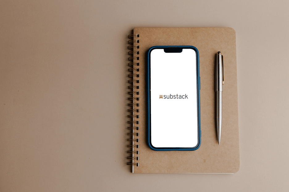 a brown notebook sitting on a brown desk with a cellphone on the notebook with the word Substack lit up on it.