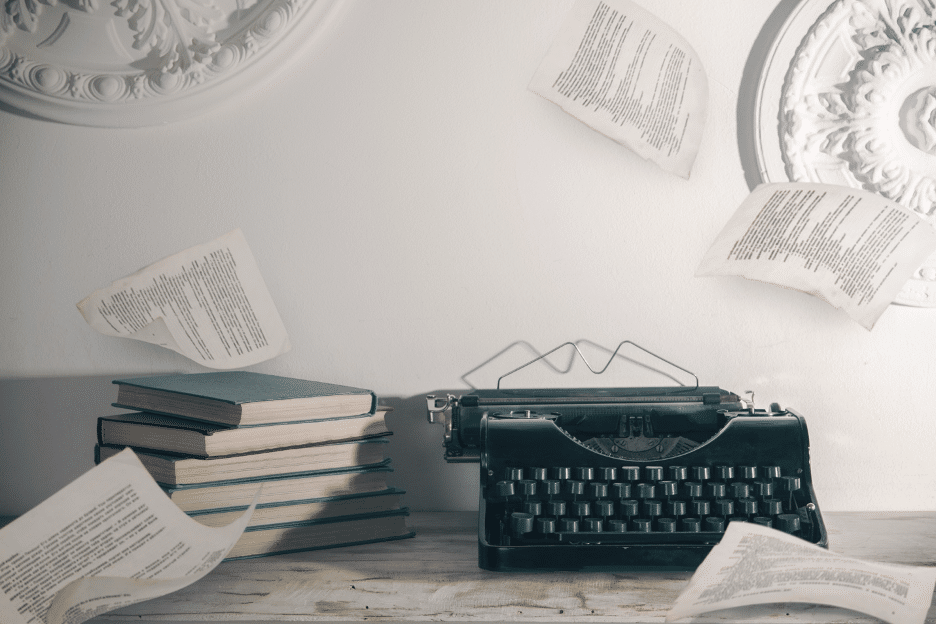 A typewriter and a stack of books on a desk with papers flying around it.