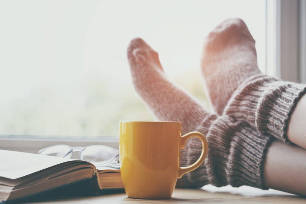 A pair of feet propped up in the sunlight beside a mug and book.