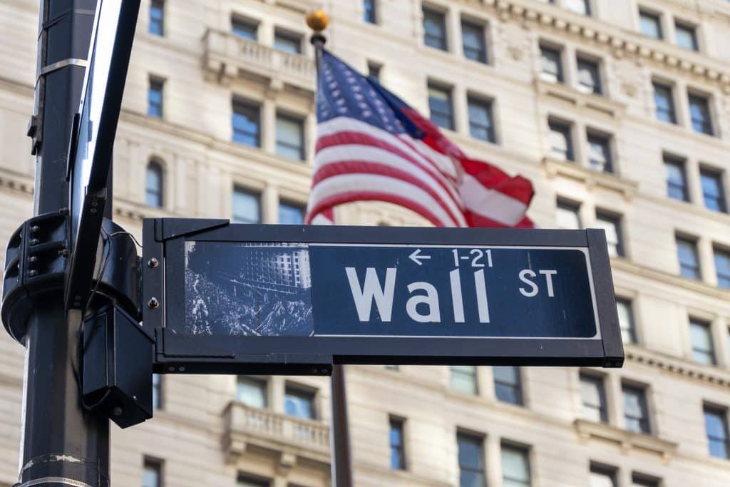 Wall St sign with American flag behind and large building complex as background.