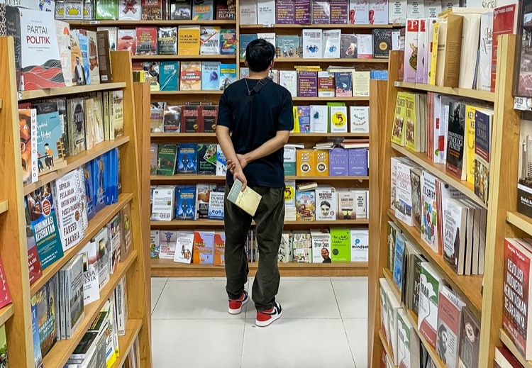A man with his back turned looks with his hands behind his back at a shelf in a bookstore.