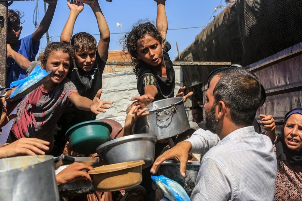 Image shown Palestinian children surrounding an older man passing out food in a camp in Palestine during the war.