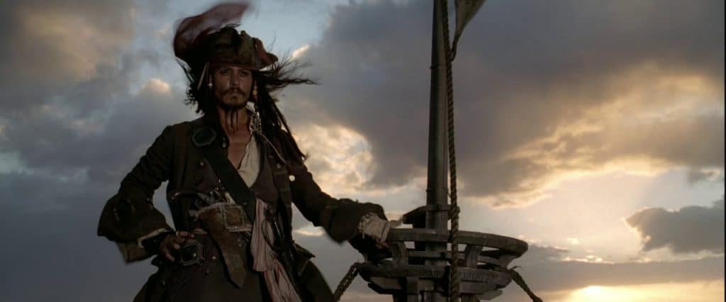 The image is a still from the first Pirates of the Caribbean movie featuring Johnny Depp as Captain Jack Sparrow standing on a ship.