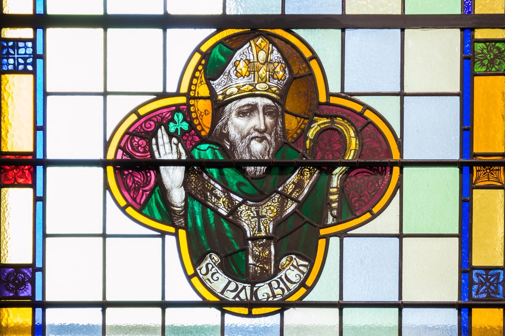 A stained glass image of Saint Patrick dressed in green and posing with a shamrock.