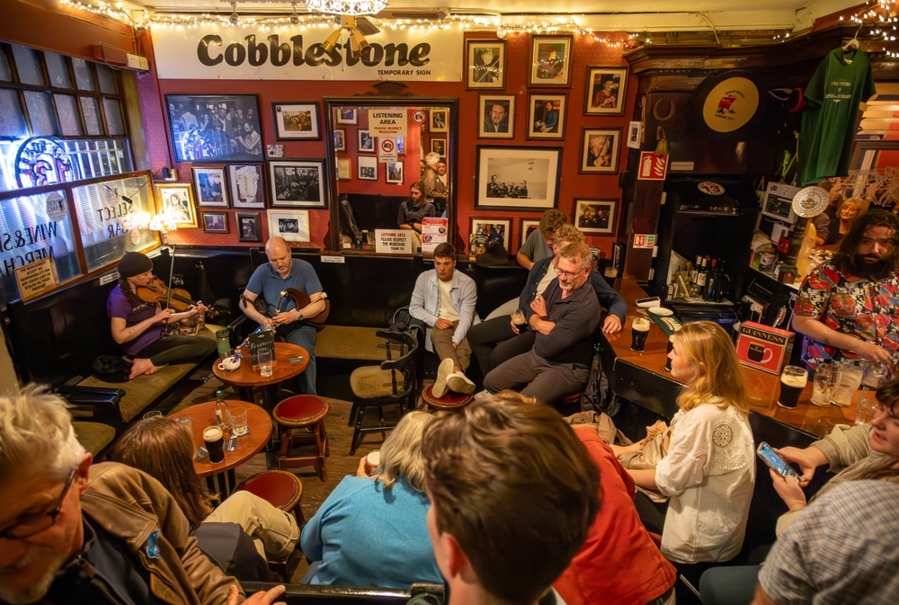 A group of people gather around the crowded Irish pub all focused on the musicians and drinking Guinness