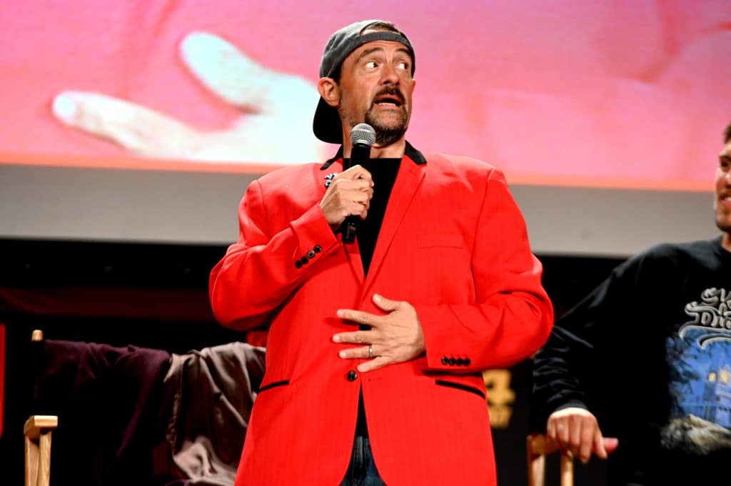 Kevin Smith stands with a microphone in hand, addressing an audience. He is wearing a bright red blazer.