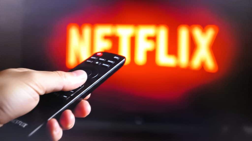 A person holds a remote in the image's forefront. In the background, the Netflix logo shows on a television