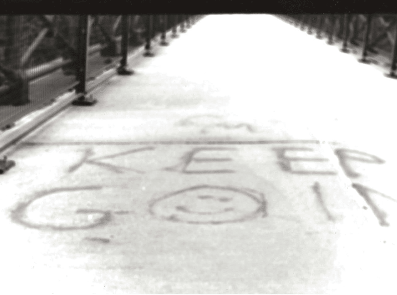 Black and white film photograph of a bridge that has "Keep Going" written on it, with the "O" being a smiley face