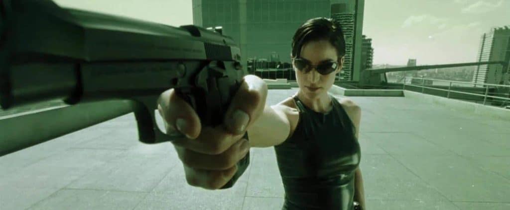 The image is a still from Matrix, featuring the character Trinity standing outside aiming a gun.