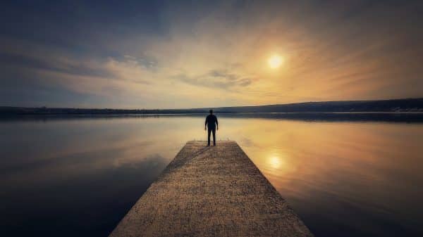 Man stands alone on the end on a dock of a lake and watches the sun's reflection in the water.