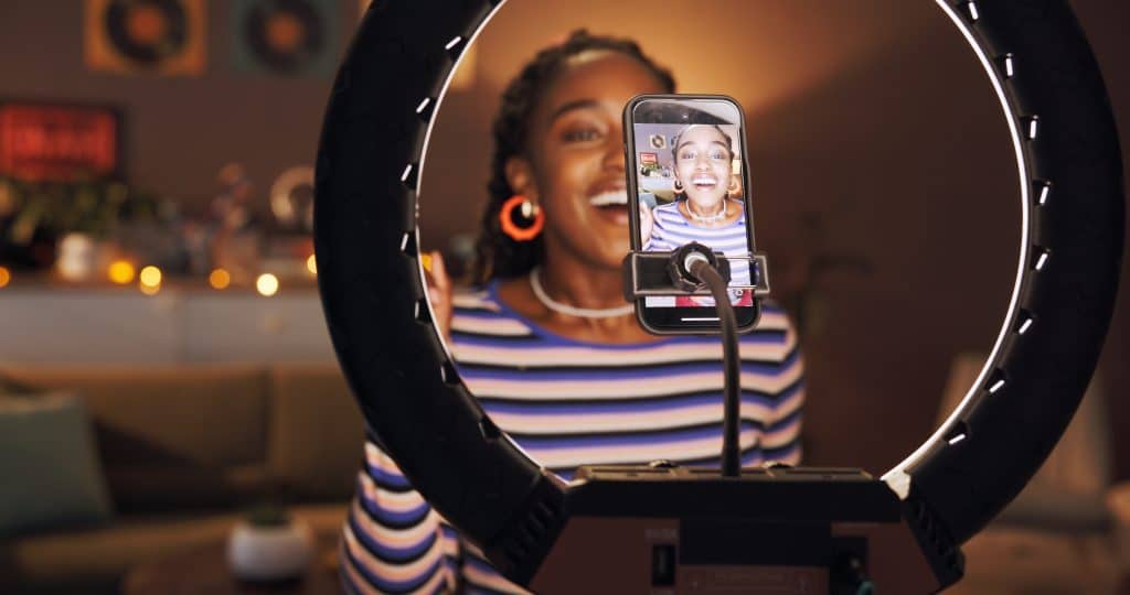 Woman sits in front of phone camera and ring light filming herself for social media.