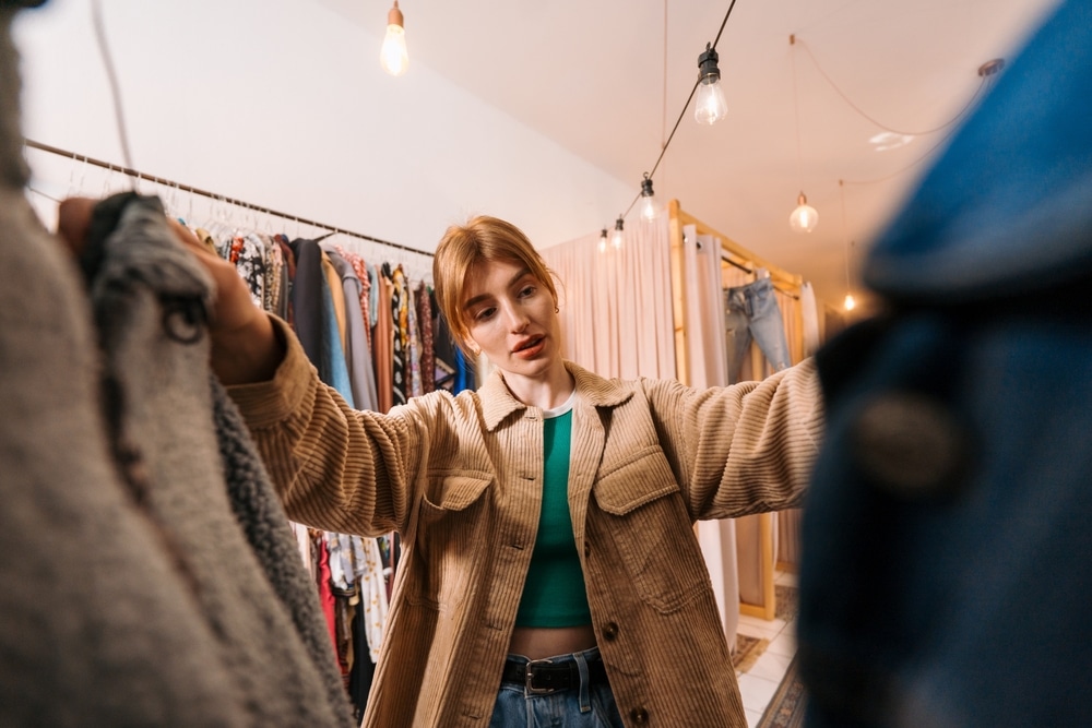 A person stands in a clothing store, looking through clothes while holding two pieces of clothing apart to examine them.