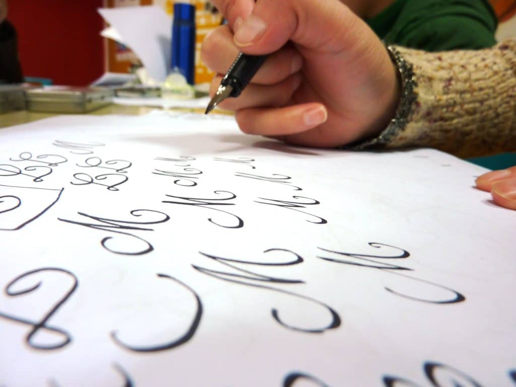 A hand with a fountain pen depicted above a paper filled with cursive letter M.