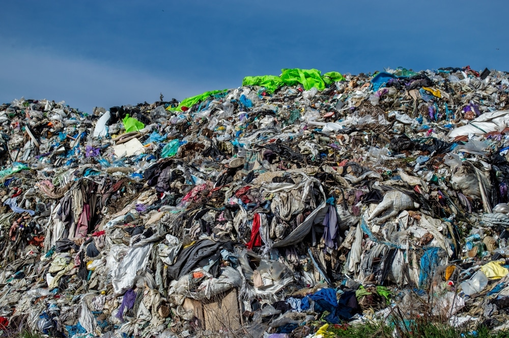 A vast heap of various discarded textiles rises against a bright blue sky, showcasing the overwhelming issue of textile waste in landfills