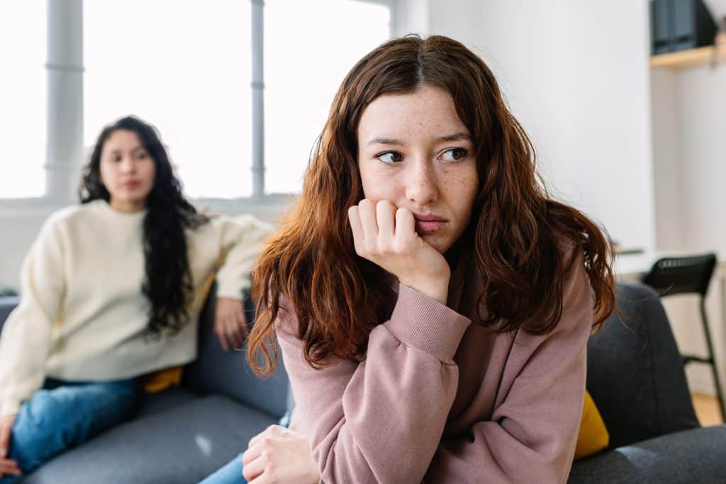 One girl looking away from her friend, visibly upset. The friend sits in the background.