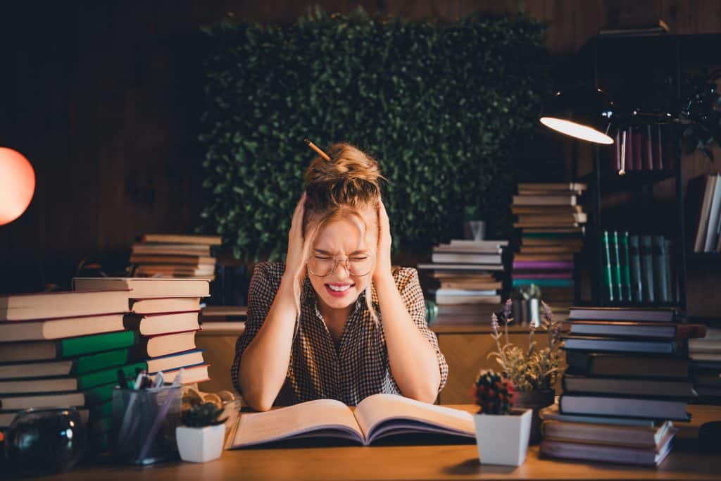 A stressed female student sits at a desk surrounded by stacks of books, holding her head while studying late at night under a desk lamp.
