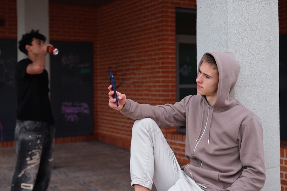 Teen boy looking at his phone while appearing emotionally detached from others nearby.