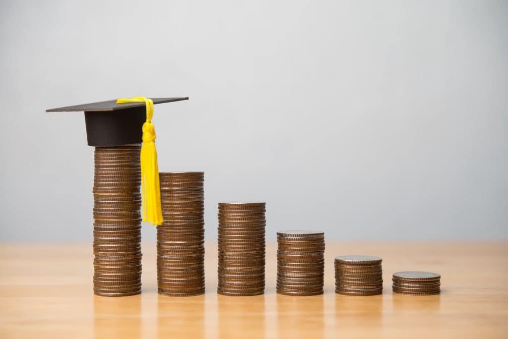 Mortarboard or square academic cap and reduce stack coins chart on wooden table with white wall background. Private student loan, student debt interest rates fall, relief repayment, education concept.