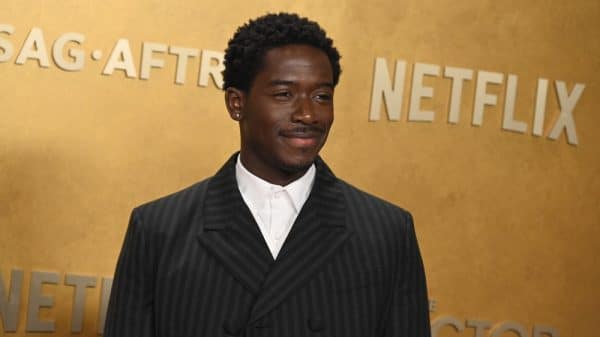 Damson Idris stands smiling in front of a gold backdrop for formal red carpet event.