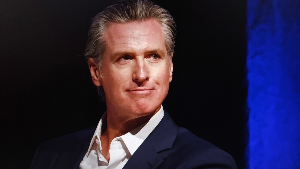 Gavin Newsom smiles slightly in a dimly lit setting, wearing a suit jacket, his hair neatly styled.