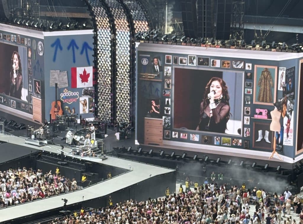 Shania Twain a woman in her 60s with long brown hair in a black leather dress, is on a big screen inside of a picture frame with multiple ones around het