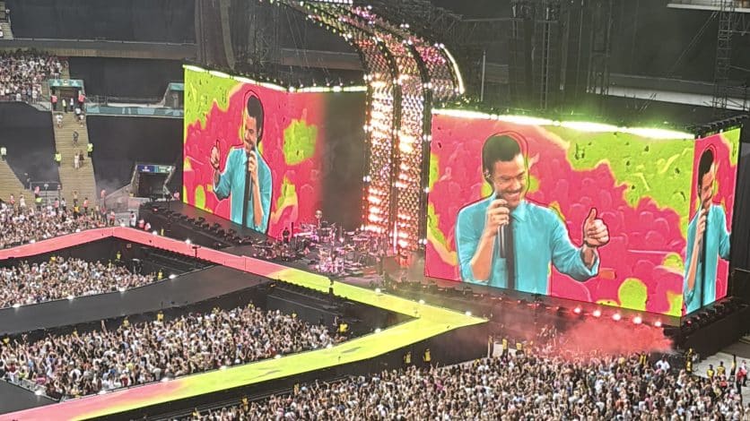A big screen with a pink and green background with harry styles smiling in front of the colours