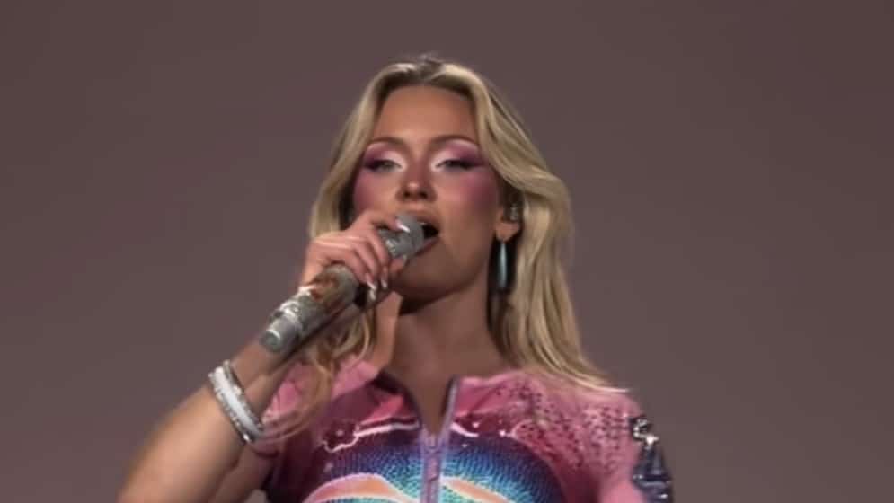 Zara Larsson sings into a glittering microphone while wearing pink makeup and a colorful performance outfit.