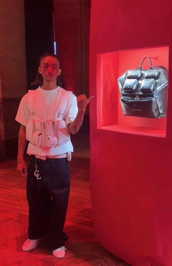 Jaden Smith, face painted red, stands in front of a display for one his bags which is a black travelling backpack with many segmented pockets.