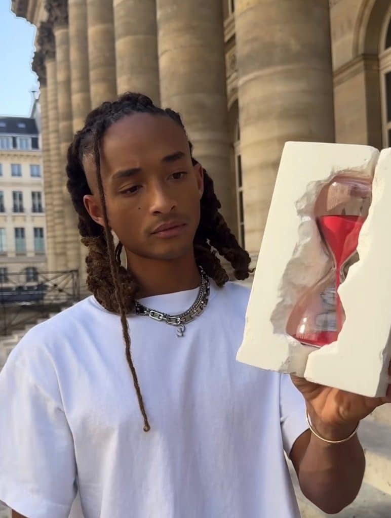 Jaden Smith poses with a red hour glass, which sits between a pale marble brick. This is one of the invitation for his collection.
