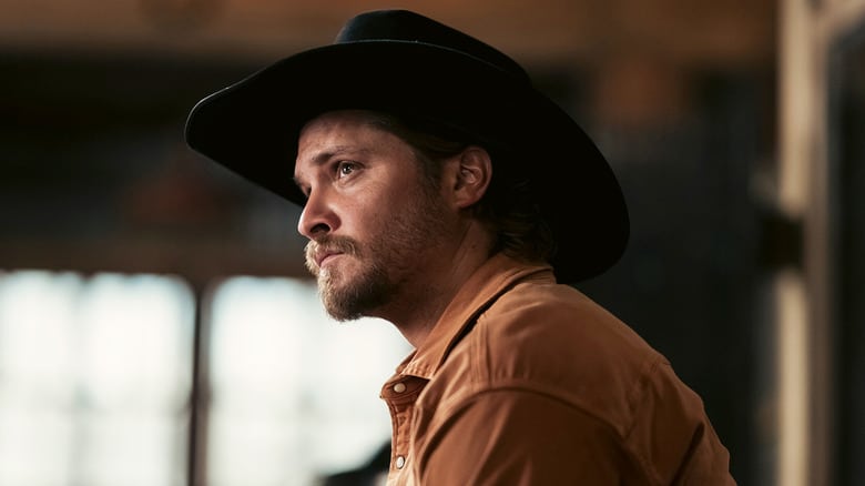 A still from Marshals where Luke Grimes is looking away from the camera in a black cowboy hat and brown jacket