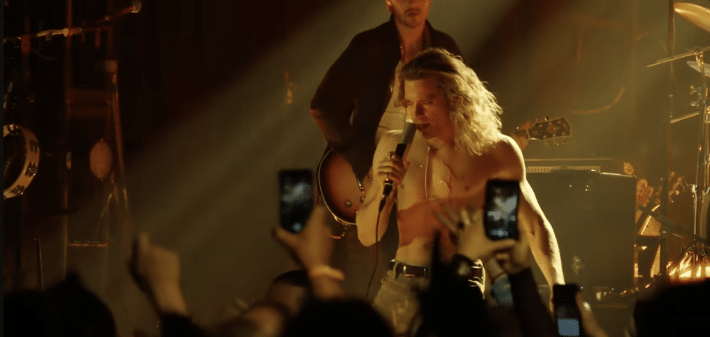 The Vampire Lestat in the midst of one of his concerts, staring haughtily at the audience while gripping a mic. He sports scars and is covered in glitter.