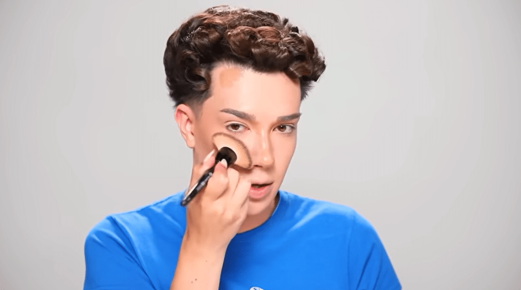 James Charles holds a brush to his face mid-makeup look.