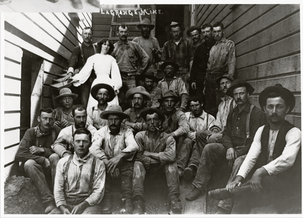Historic black-and-white photograph of a large group of Gold Rush-era miners and laborers gathered on wooden stairs outside a mine building. The men wear work clothes, suspenders, hats, and boots, while a woman in a white dress stands near the center. The image reflects the rugged working conditions and communities associated with late nineteenth-century American mining culture.