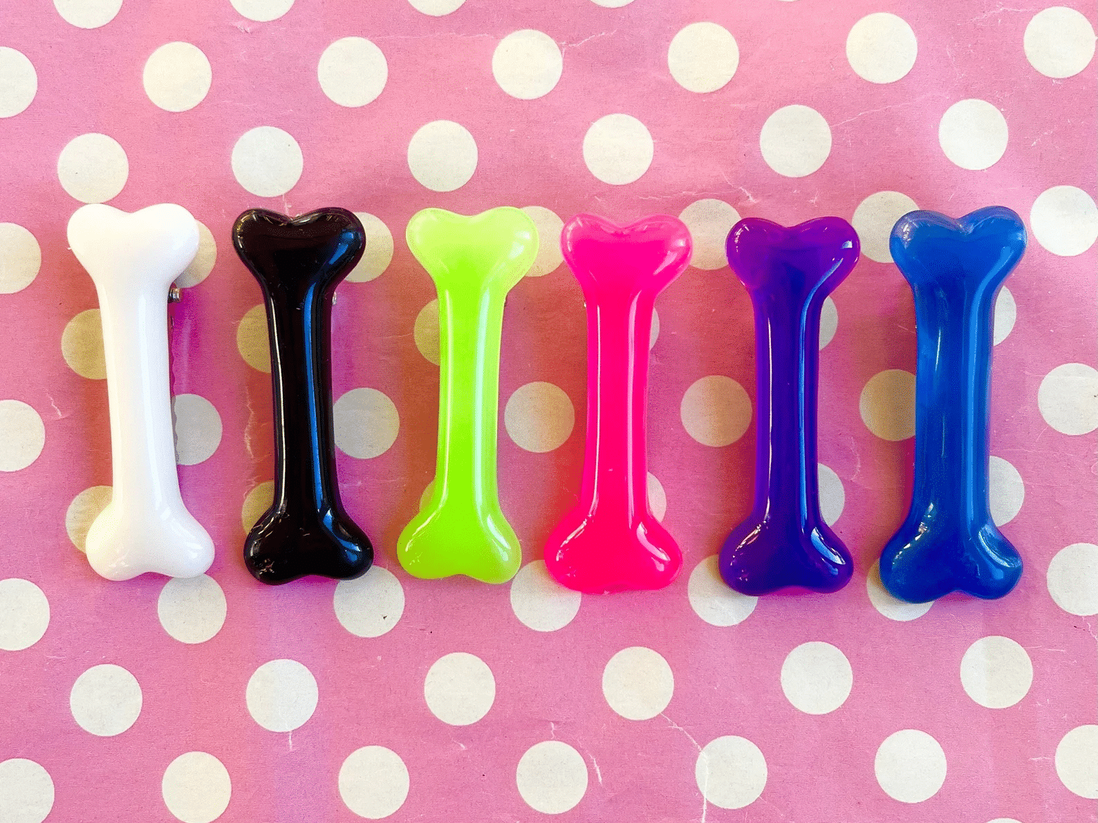 All of KawaiiKave's bone clip colors (from left to right: white, black, green, pink, purple, blue) are aligned horizontally against and a pink background that has white polka dots.