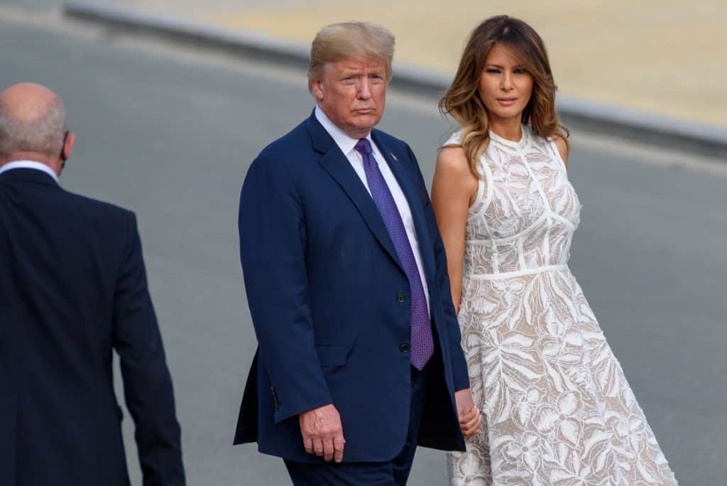 President Trump and Melani stand close together. He is wearing a blue suit and she is wearing a white lace dress.