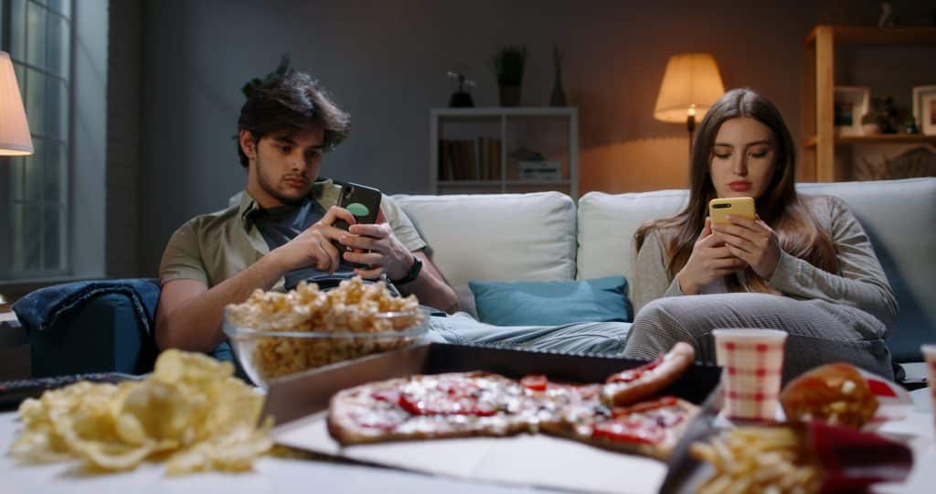 A man and a woman sit looking at their phones while various movie-watching objects lie on the table in front of them