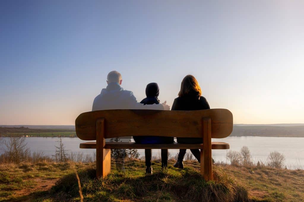 Two women sit on a bench looking out at a river, a faded man puts his arm over the shoulder of one of the women, meant to symbolise the grief of these two women and the loss of the man