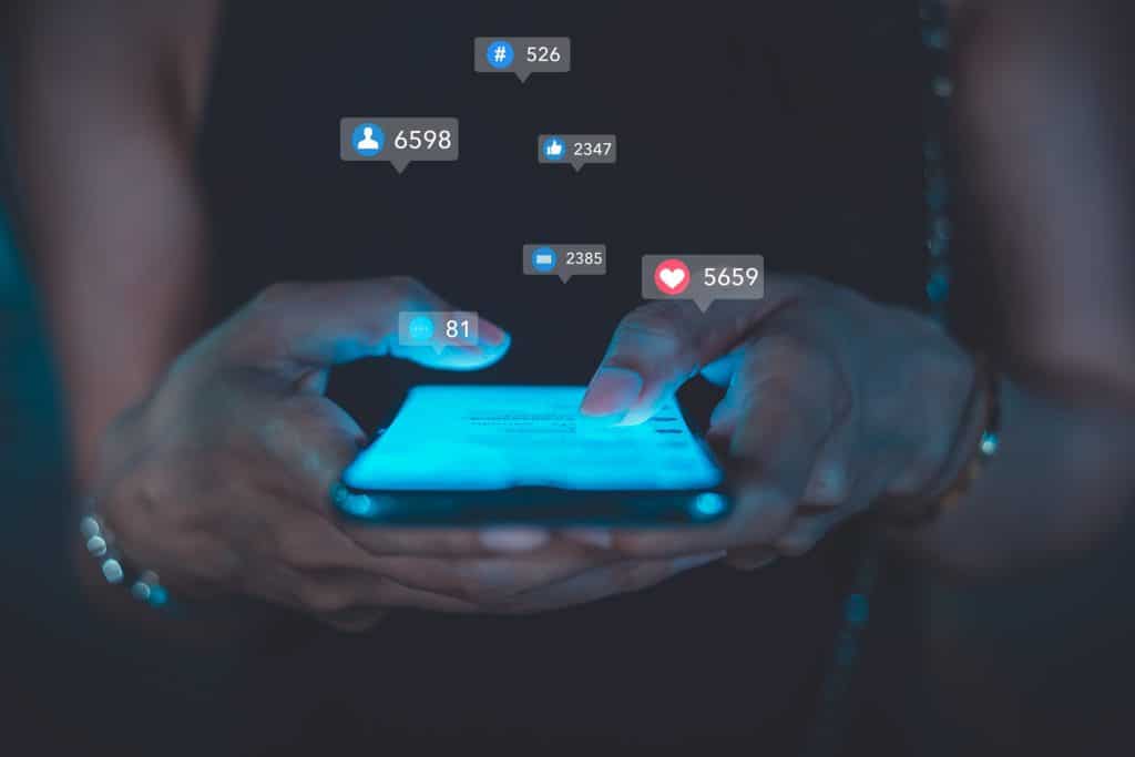 Two hands are gripping a phone. Blue light emits from the phone, and various social media icons are popping out from the device.