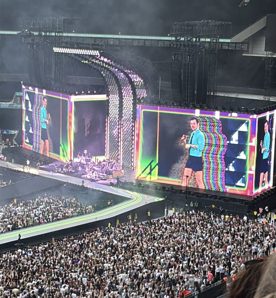 Harry Styles 32 year old man with brown hair, blue shirt on a screen in Wembley Stadium, with multiple colours like pink, green, yellow and blue surrounding him
