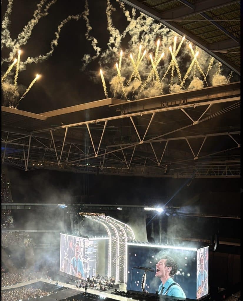 Harry Styles 32 year old man with brown hair, blue shirt on a screen in Wembley Stadium singing with fireworks coming out the top of the stadium roof